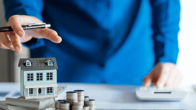 A person in a blue shirt reaches for a miniature house and coins on a desk