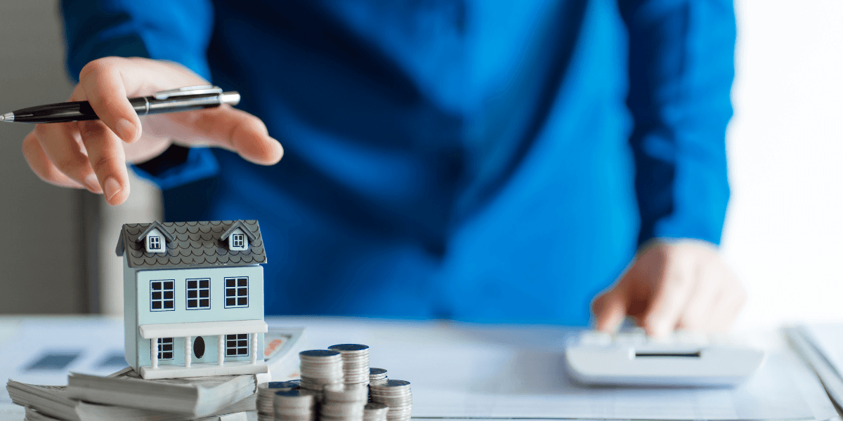A person in a blue shirt reaches for a miniature house and coins on a desk