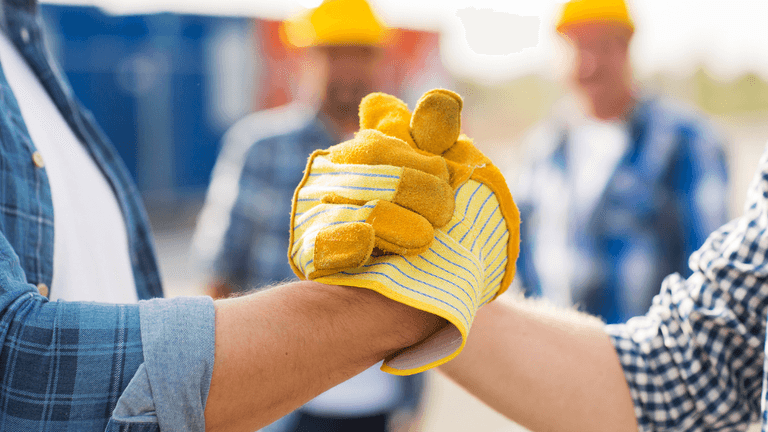 Two people with gloves clasping hands, construction workers in the background.