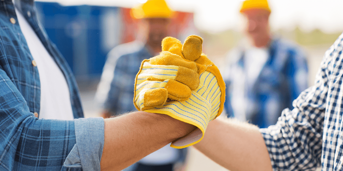 Two people with gloves clasping hands, construction workers in the background.