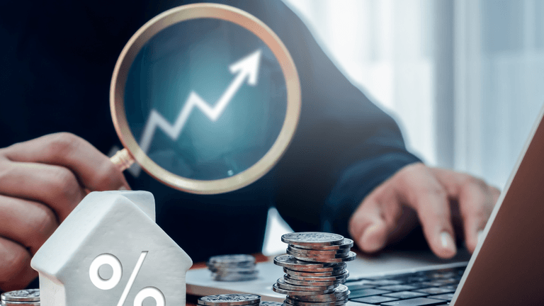 Person examining a rising graph with a magnifying glass, coins and a house figure nearby