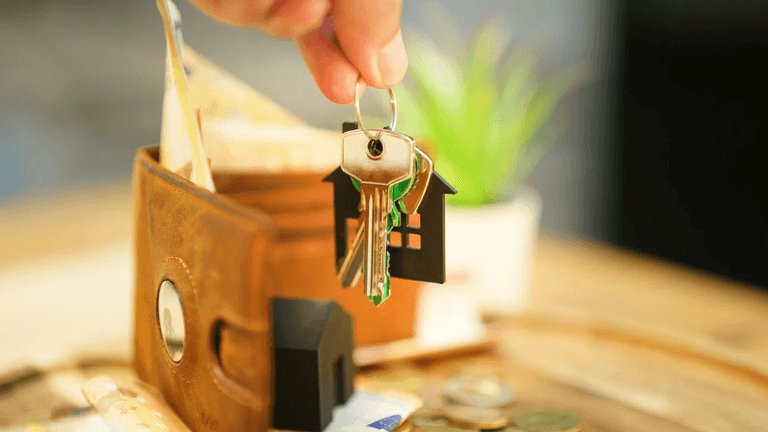 A hand holding house-shaped key over a wallet with cash and coins