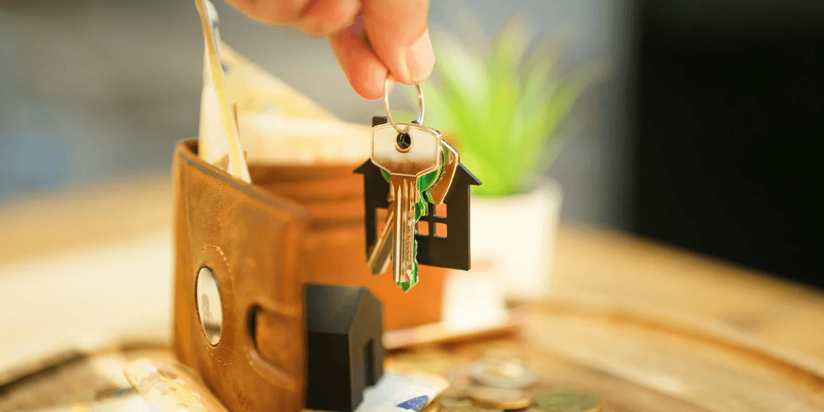 A hand holding house-shaped key over a wallet with cash and coins