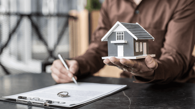 Person holding a model house while signing real estate paperwork at a desk.